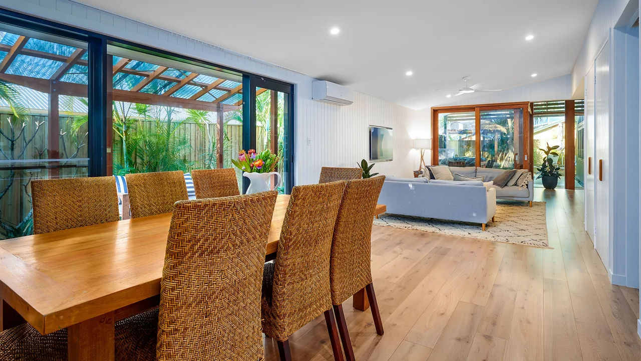 A dining table under a glazed wall, looking the length of a timber-lined house past the kitchen to the living room.
