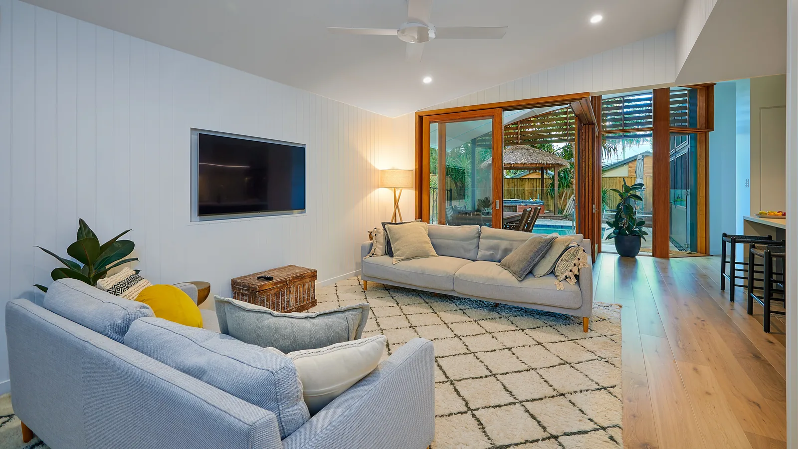 A living room lined in vertical white boards, opening through timber-framed sliding doors and louvres to a pool terrace, with oak flooring running through to the kitchen.
