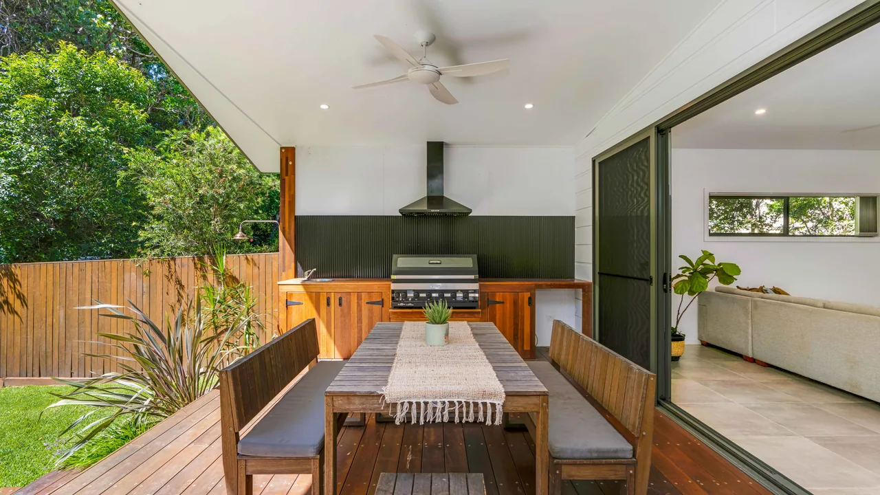 A covered outdoor room off the back of a house, with a hardwood table and a built-in barbecue under a rangehood.