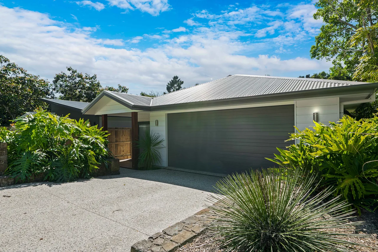 A single-storey home seen from its driveway. Pale weatherboard cladding and a dark panelled garage door under a low-pitched Colorbond roof, a timber gate and hardwood posts at the entry, subtropical planting to both sides and an exposed-aggregate drive in front.