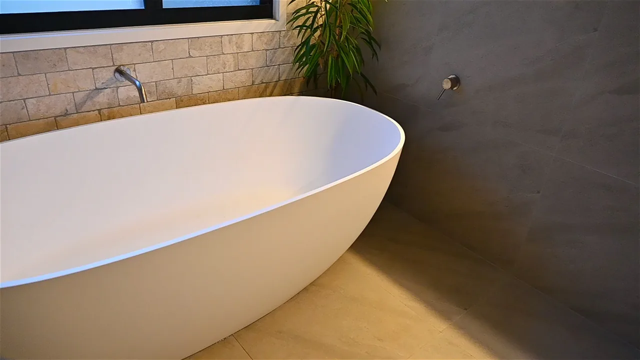 A freestanding oval bath in matte white, set into the corner of a bathroom. Behind it a travertine-tiled splashback with a wall-mounted spout, and to the side a large-format concrete-look wall with a round wall mixer.