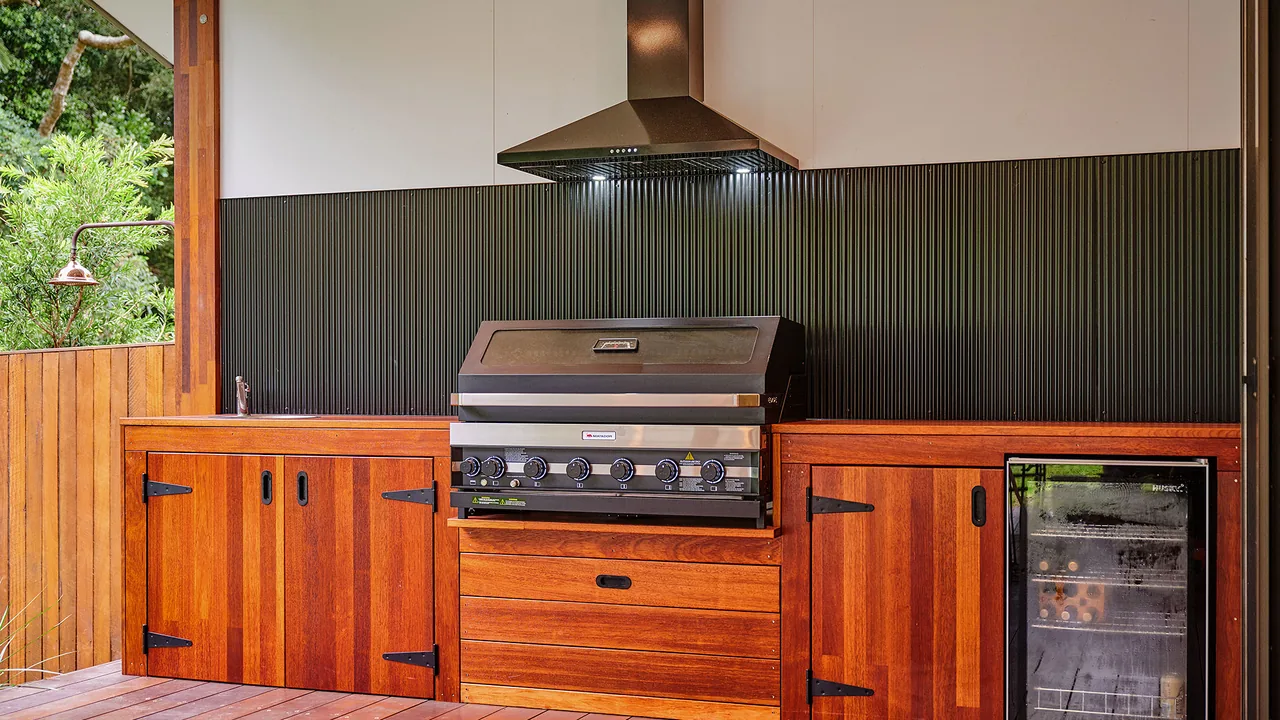 An outdoor kitchen built into hardwood joinery on a hardwood deck, with a stainless rangehood and a dark corrugated splashback.