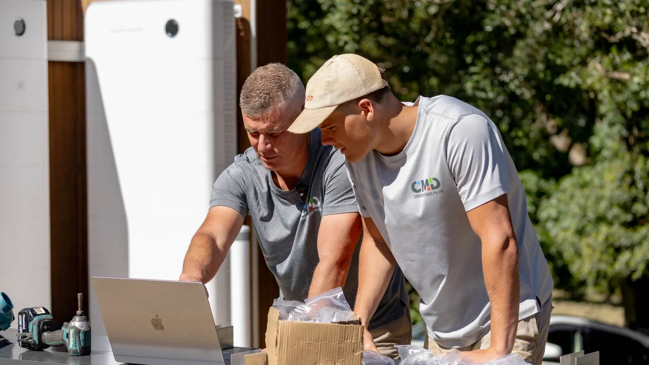 Cade Davidson and a member of his team leaning over a laptop on a bench on site, working something out, with cordless tools and packaging beside them.