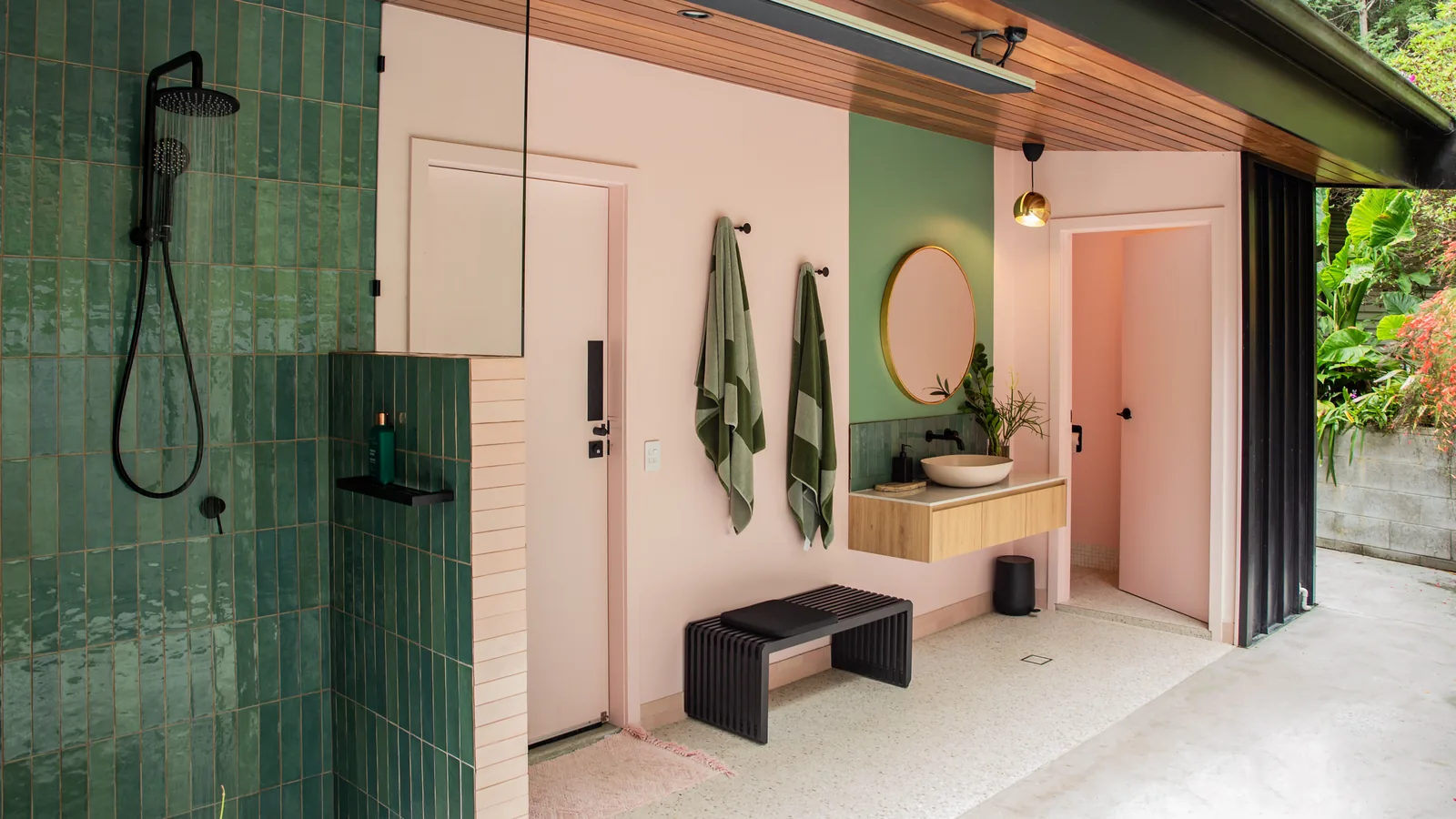 The open-air bathroom: green tiled shower, pink walls, a round mirror over a basin, and the far end open to the garden.