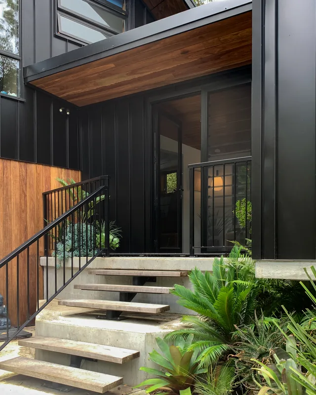 Concrete entry steps rising between planting to a black-clad wall and a glazed entry door.