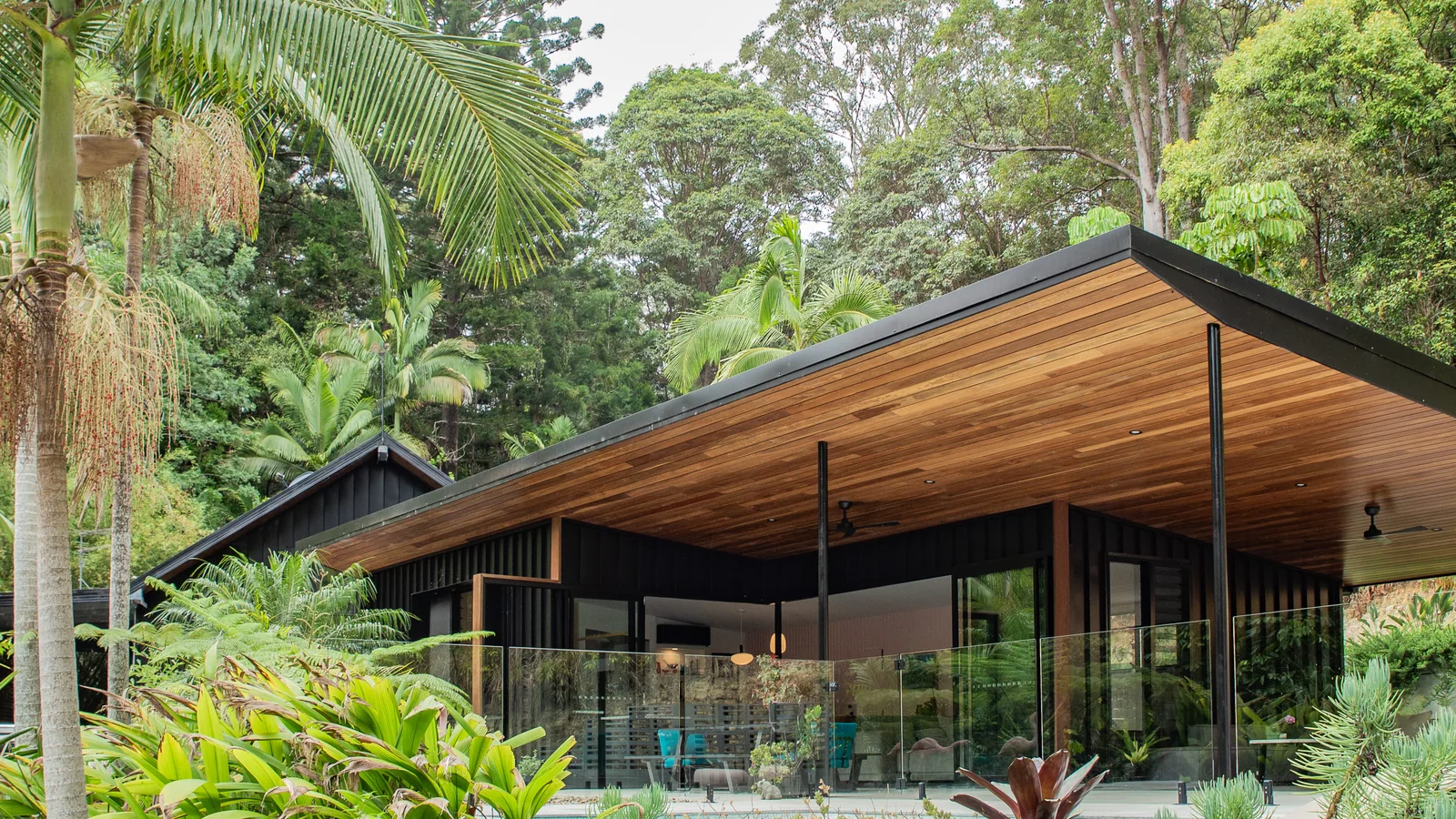 A long low house with a raking timber-lined roof, seen from the driveway through palms and subtropical planting.