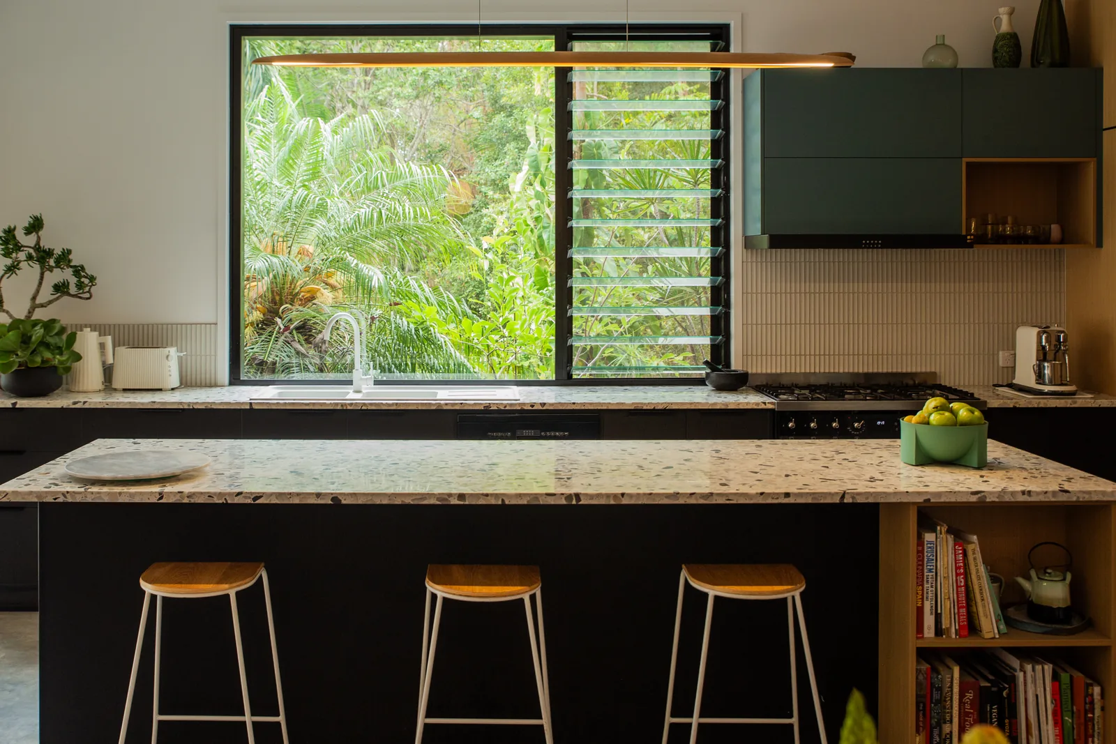 A kitchen with a dark island and stone benchtop, green upper joinery, and a louvred window over the sink looking into the garden.