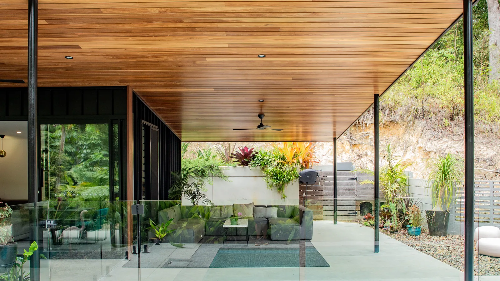 A covered outdoor room lined in spotted gum, open on three sides, with a lounge, a plunge pool and a planted wall beyond.