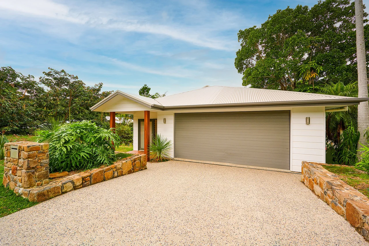 A new single-storey home seen from the street. Pale weatherboard cladding under a hipped Colorbond roof, a double garage, and a covered entry porch on a hardwood post. Stone retaining walls run along both sides of a wide exposed-aggregate driveway, with subtropical planting and mature trees behind.