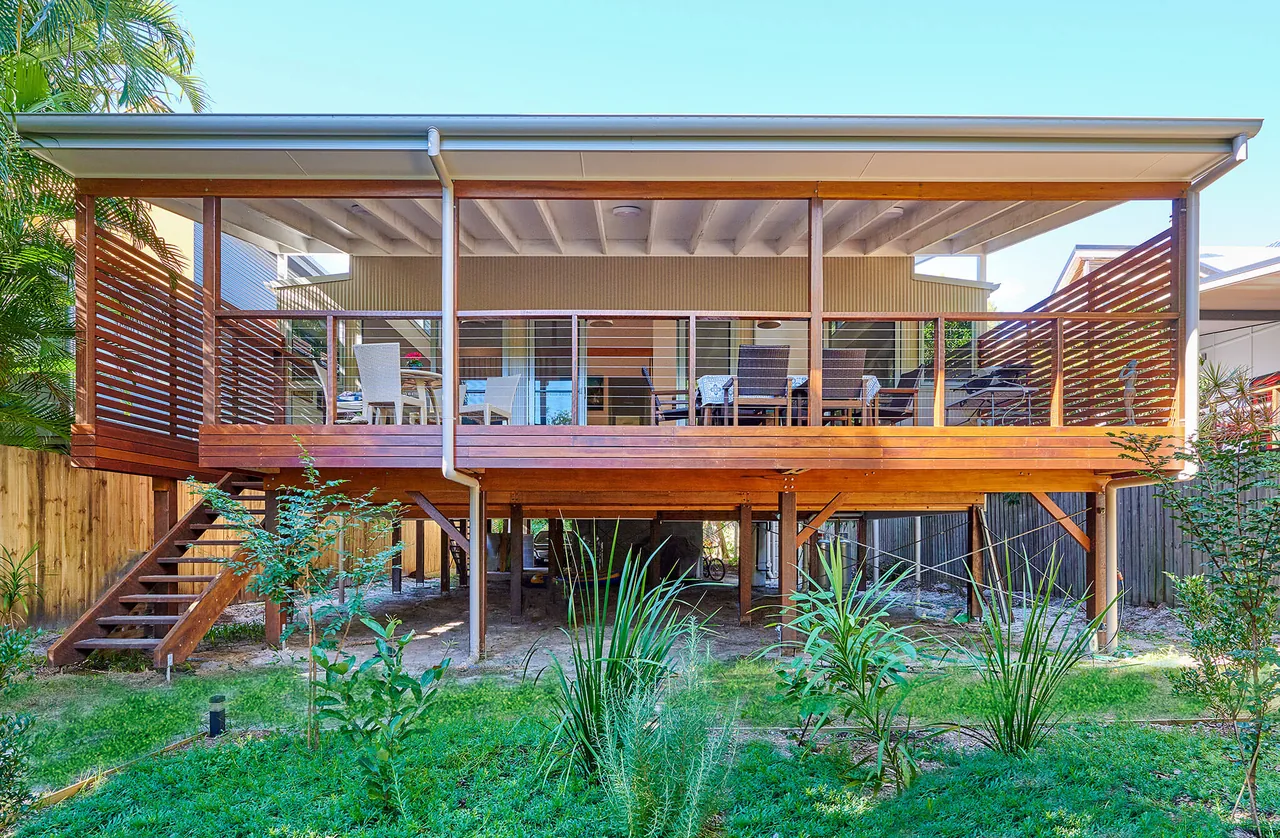 A raised timber deck across the rear of a single-storey home. Hardwood posts and beams carry a flat roof over the deck, with horizontal slat screens at both ends, a stainless wire balustrade and a timber handrail. Sliding glass doors open from the living area onto an outdoor setting, and timber stairs run down to a newly planted garden.